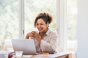Using her laptop and bluetooth earbuds, the mid adult woman video conferences from home during the coronavirus epidemic.